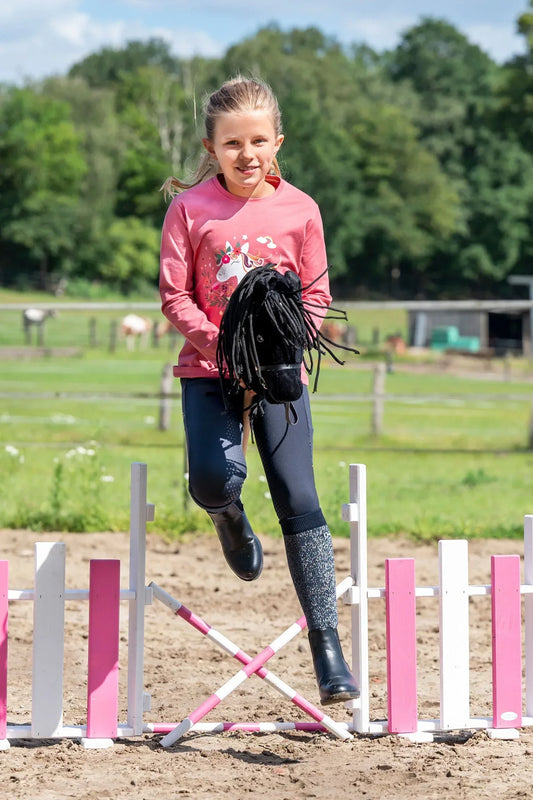 Blondes Mädchen spring mit schwarzem Hobby Horse über ein Hindernis