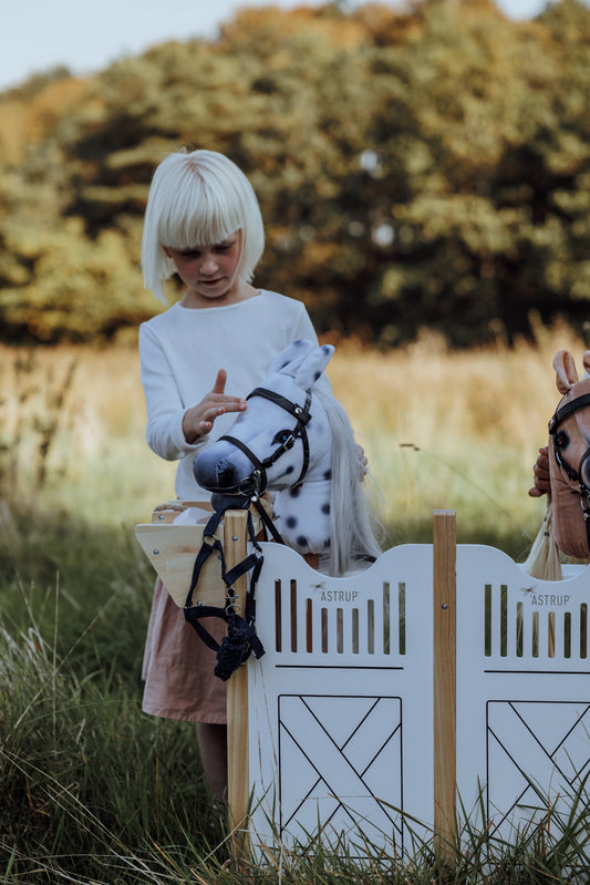 Kind pflegt ihr plüschiges Hobby Horse an dem stilvollen weißen Hobby horse Stall aus Holz im skandinavischen Design.