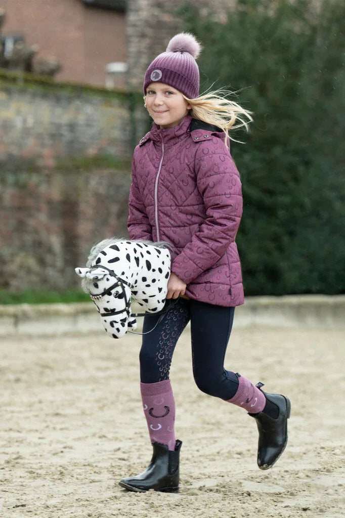 HKM Hobby Horse Knapstrupper wird von einem blonden Mädchen über einen Reitplatz galoppiert. Das Mädchen trägt HKM Reitkleidung in lila.