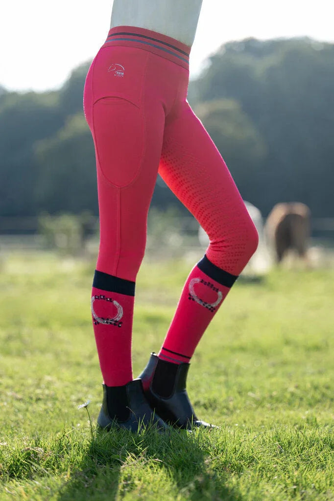 HKM Reitleggings mit Vollbesatz aus Silikon und Kniestrümpfe in Pink; Detailansicht der Reiterbeine vor einer grünen Pferdeweide