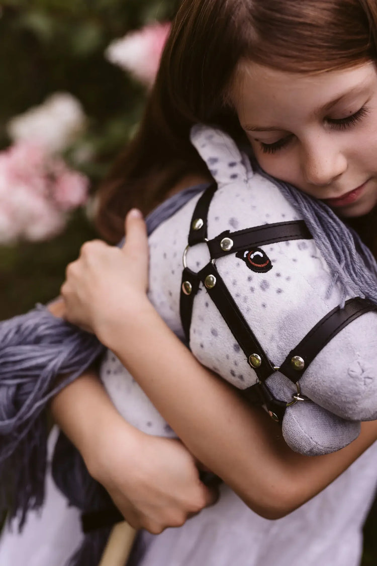 Aufnahme eines Mädchens, dass sein Hobby Horse im Arm hält. Das Hobby Horse ist weiß mit grauer Mähne und trägt eine schwarze Hobby Horse Trense.