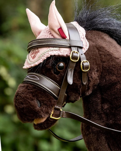 Realistisches Steckenpferd-Zubehör für Turniere in Deutschland: Ein braunes Hobby Horse mit rosa Fliegenhaube trägt die braune LeMieux Turniertrense aus Leder beim Sprung- oder Dressurtraining.