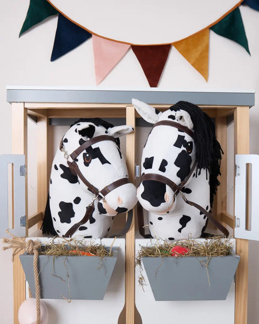 Zwei Bieco Hobby Horses in einem Hobby Horsing Stall unter einr Wimpelkette, vor ihnen Heu in einem Futtertrog. Die Hobby Horses entsprechen der Größen M+L, sind schwarz-weiß gescheckt und tragen braune Trensen.