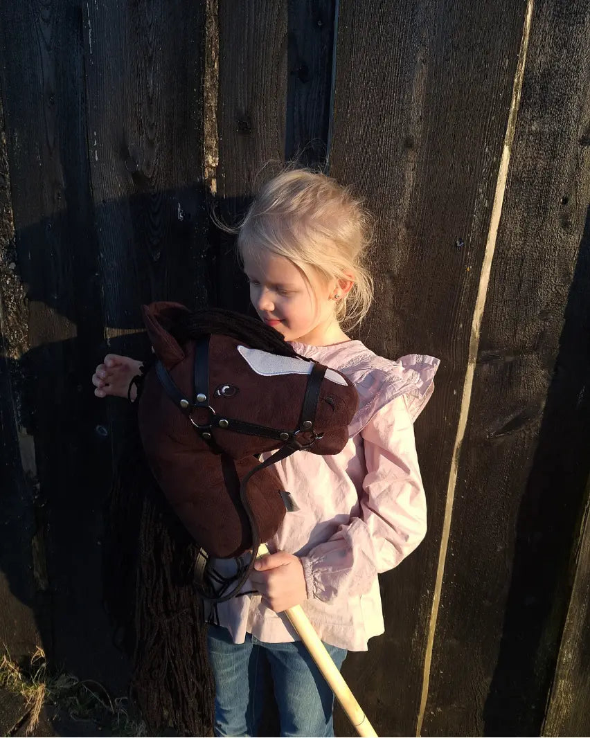 Ein Kind hält das dunkelbraune byASTRUP Hobby Horse im Arm; authentisches Steckenpferd mit schwarzer Mähne und Zaumzeug vor einer rustikalen Holzwand