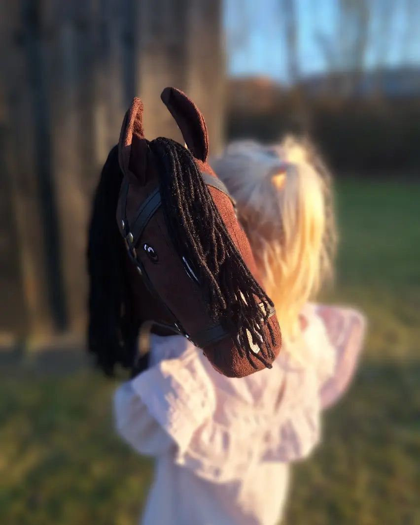 Nahaufnahme des byASTRUP Hobby Horses 'Braun' im Sonnenlicht auf einer Wiese auf dem Arm eines blonden Mädchens; weicher Stoff und realistische Augenpartie mit Trense.