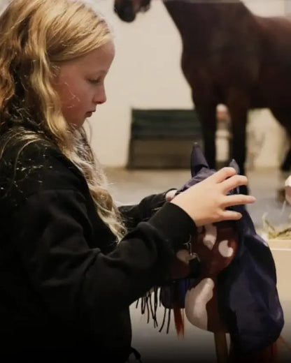 Ein Kind zieht einem byASTRUP Hobby Horse einen dunkelblauen Regenschutz über; authentisches Zubehör für das Spiel im Stall.