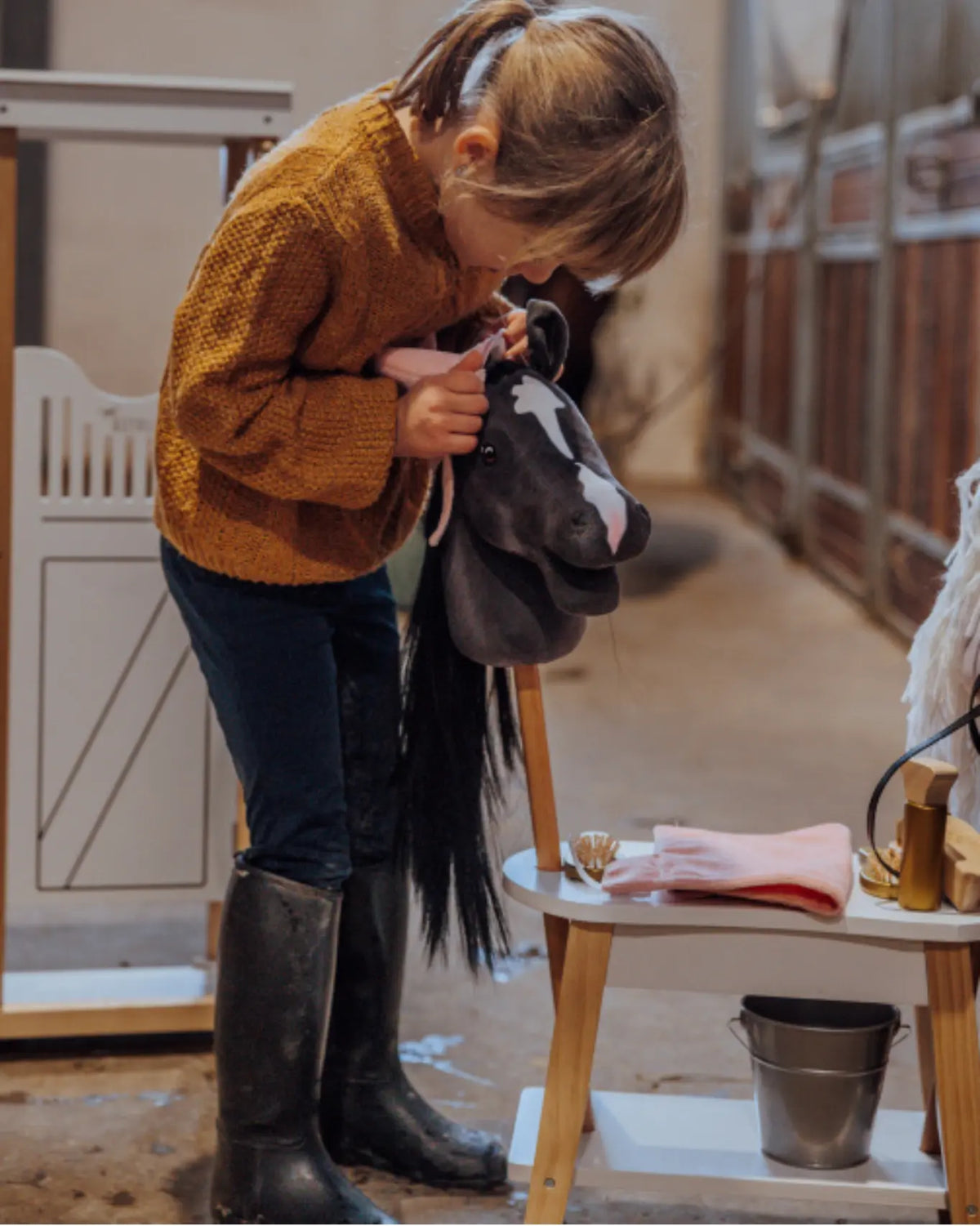 Mädchen bereitet ihr Hobby Horse im Stall vor; altrosafarbene byASTRUP Decke und Bürste auf der Holzbank.