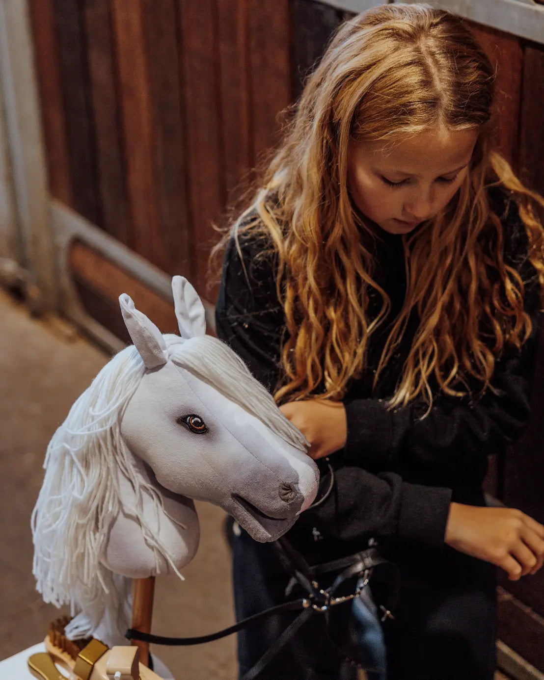 Kreative Pferdepflege beim Hobby Horsing: Ein Mädchen frisiert und flechtet die weiche Wollmähne des weißen byASTRUP Steckenpferds an einem hölzernen Stalltor.