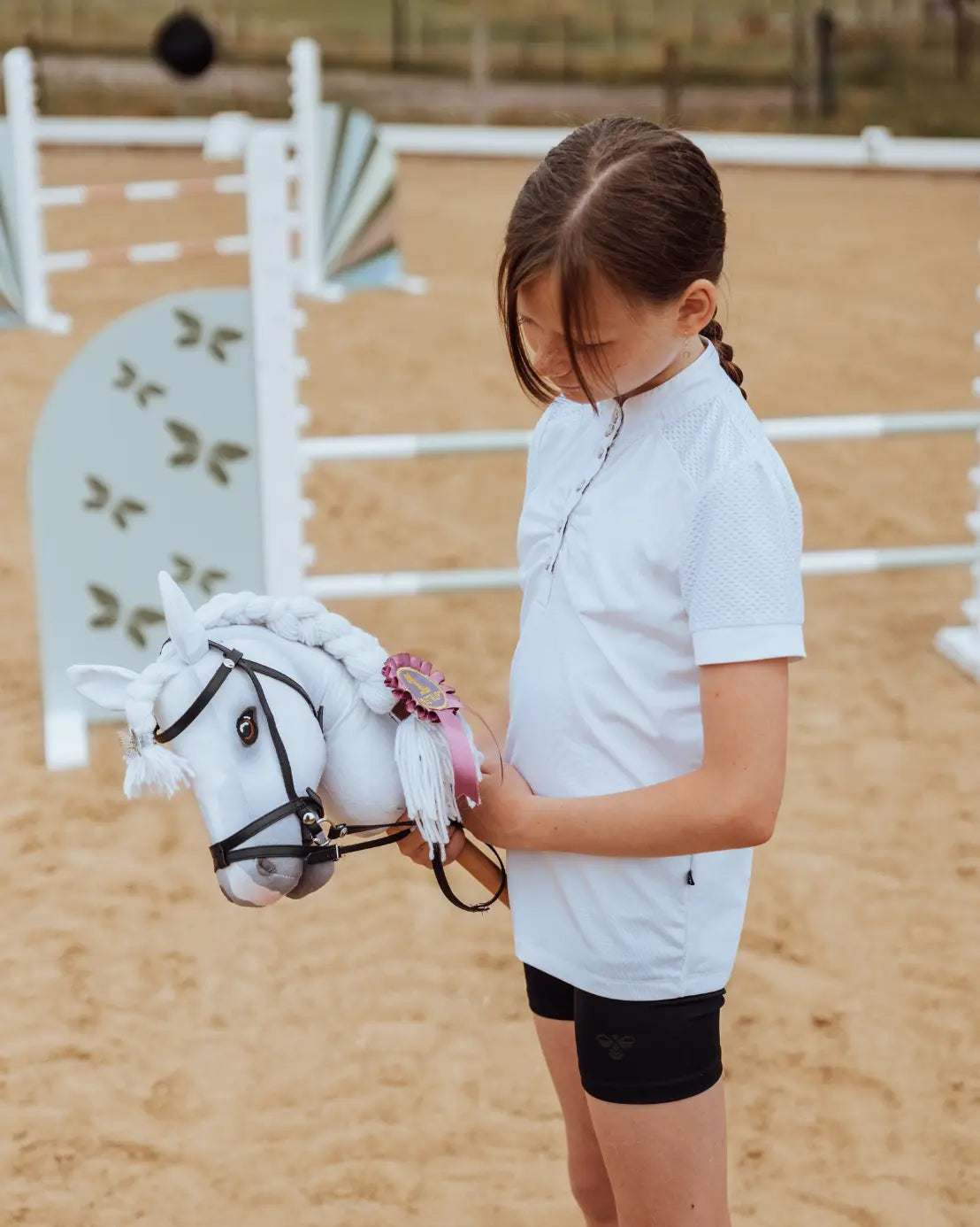 Professioneller Turnier-Look für kleine Reiter: Turnierreiterin bereitet sich mit dem weißen byASTRUP Hobby Horse Schimmel auf einem sandigen Reitplatz auf den Parcours vor.