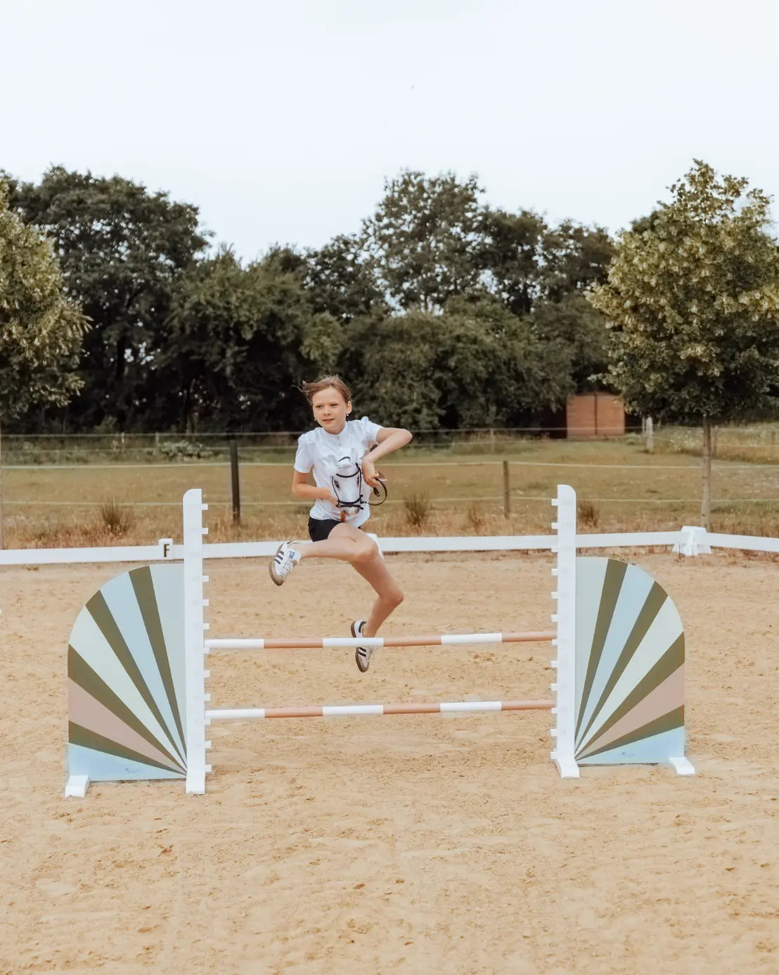 Sportliches Hobby Horsing Springtraining: Ein Kind springt barfuß mit dem leichten byASTRUP Steckenpferd über ein weiß-grünes Hindernis auf dem Turnierplatz.