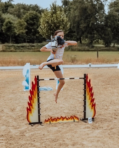 Hobby Horsing Springtraining: Ein Mädchen springt mit ihrem Steckenpferd über das feurige byASTRUP Hindernis Fire auf einem sandigen Reitplatz.