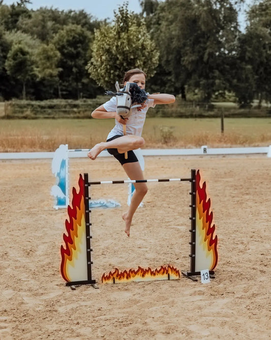 Hobby Horsing Springtraining: Ein Mädchen springt mit ihrem Steckenpferd über das feurige byASTRUP Hindernis Fire auf einem sandigen Reitplatz.