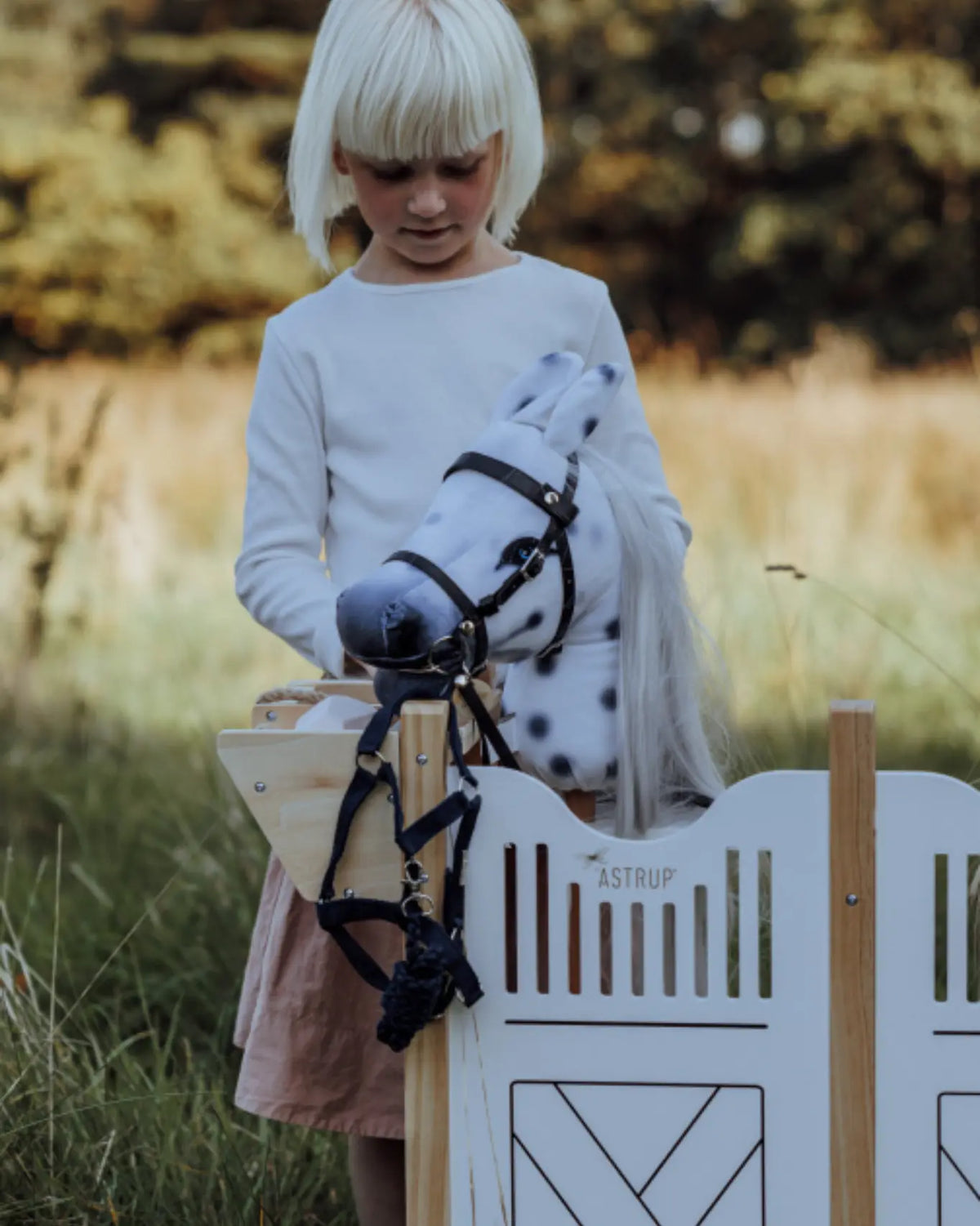 Kind steht auf der Wiese neben dem byASTRUP Holzstall; graues Hobby Horse trägt das dunkelblaue Halfter-Set.