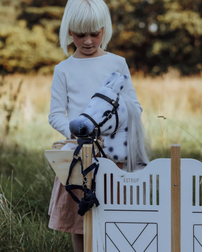 Kind steht auf der Wiese neben dem byASTRUP Holzstall; graues Hobby Horse trägt das dunkelblaue Halfter-Set.