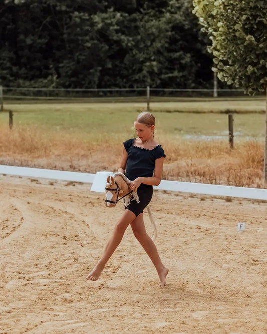 Hobby Horsing Parcours-Training: Ein Mädchen reitet barfuß mit dem beliebten byASTRUP Steckenpferd Palomino in Größe M auf einem sandigen Reitplatz.