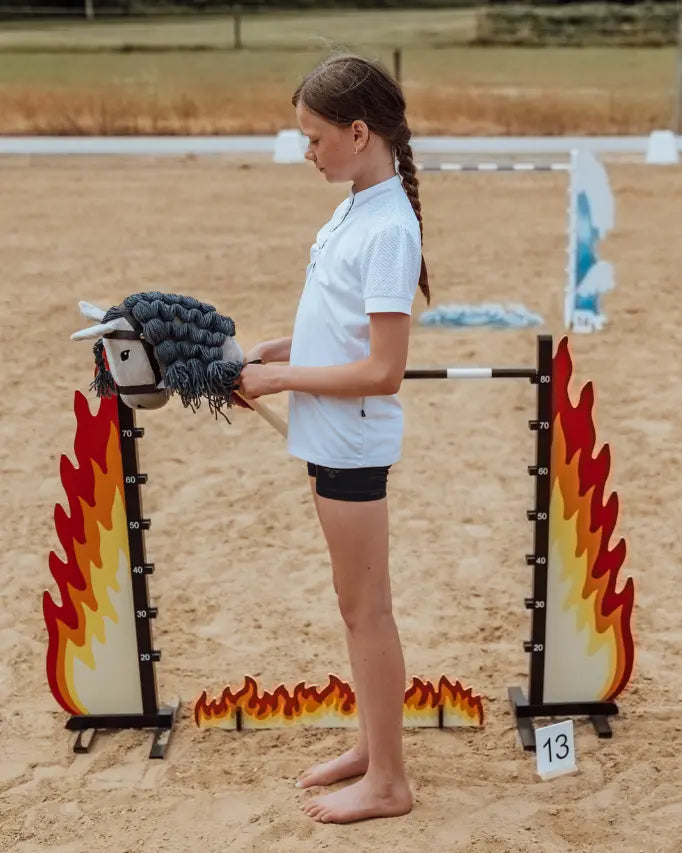 Hobby Horsing Training auf dem Reitplatz: Ein Mädchen bereitet sich mit dem grauen byASTRUP Steckenpferd auf den Sprung über ein Feuer-Hindernis vor.