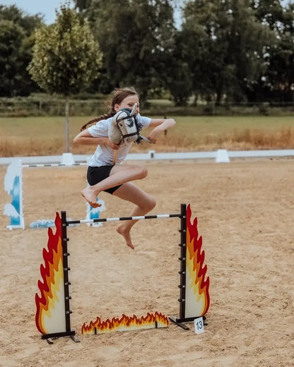 portliches Outdoor-Spiel: Dynamische Aufnahme eines Mädchens, das barfuß mit dem grauen byASTRUP Hobby Horse ein Sprunghindernis bezwingt.
