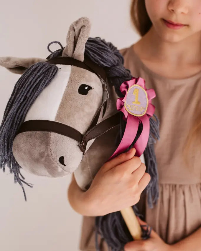Sieg beim Hobby Horsing Turnier: Ein Mädchen hält das graue byASTRUP Kuschel-Steckenpferd mit einer rosafarbenen Preisschleife stolz im Arm.