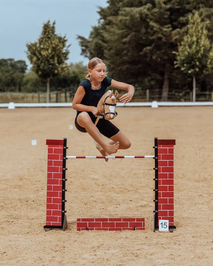Kind springt mit einem byASTRUP Hobby Horse über das rote Steinmauer-Hindernis; Hobby Horsing Training auf einem Reitplatz im Freien