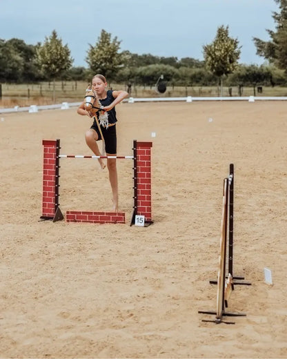 Mädchen galoppiert mit Hobby Horse über ein byASTRUP Sprunghindernis; Outdoor-Spielzeug für Hobby Horsing Sport und Bewegung
