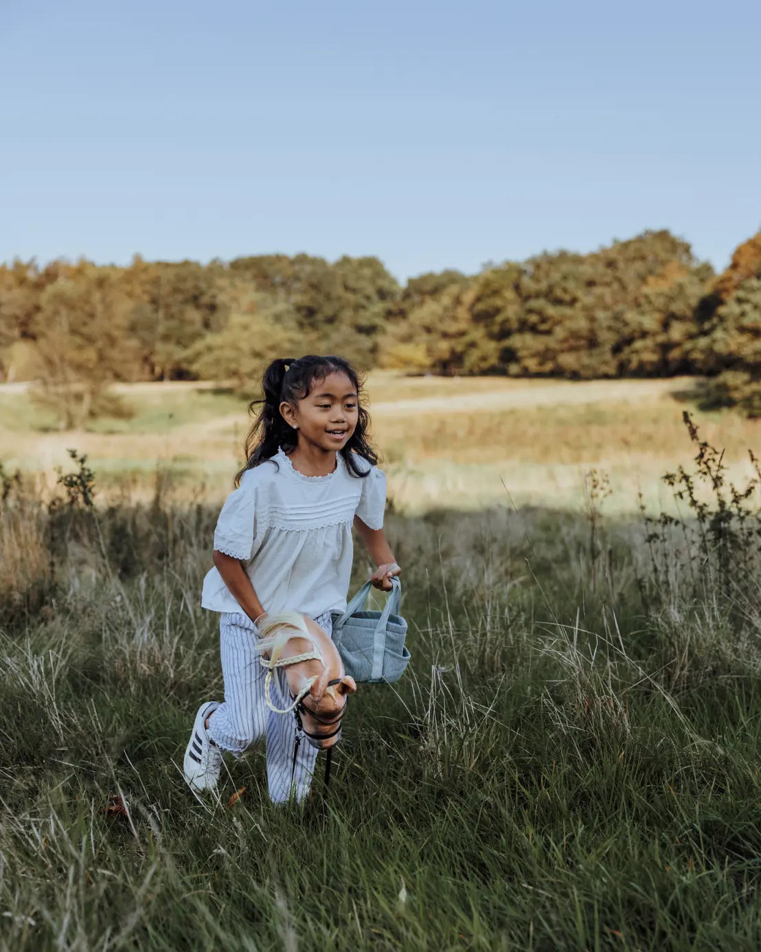 Spielspaß für unterwegs: Ein Mädchen läuft mit dem Hobby Horse und der passenden grünen byASTRUP Putztasche über eine Wiese – ideal für Outdoor-Abenteuer.