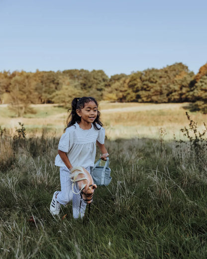 Spielspaß für unterwegs: Ein Mädchen läuft mit dem Hobby Horse und der passenden grünen byASTRUP Putztasche über eine Wiese – ideal für Outdoor-Abenteuer.