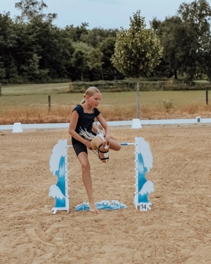 Kind trainiert Hobby Horsing auf einem Reitplatz und überspringt das blau-weiße byASTRUP Hindernis Wave.