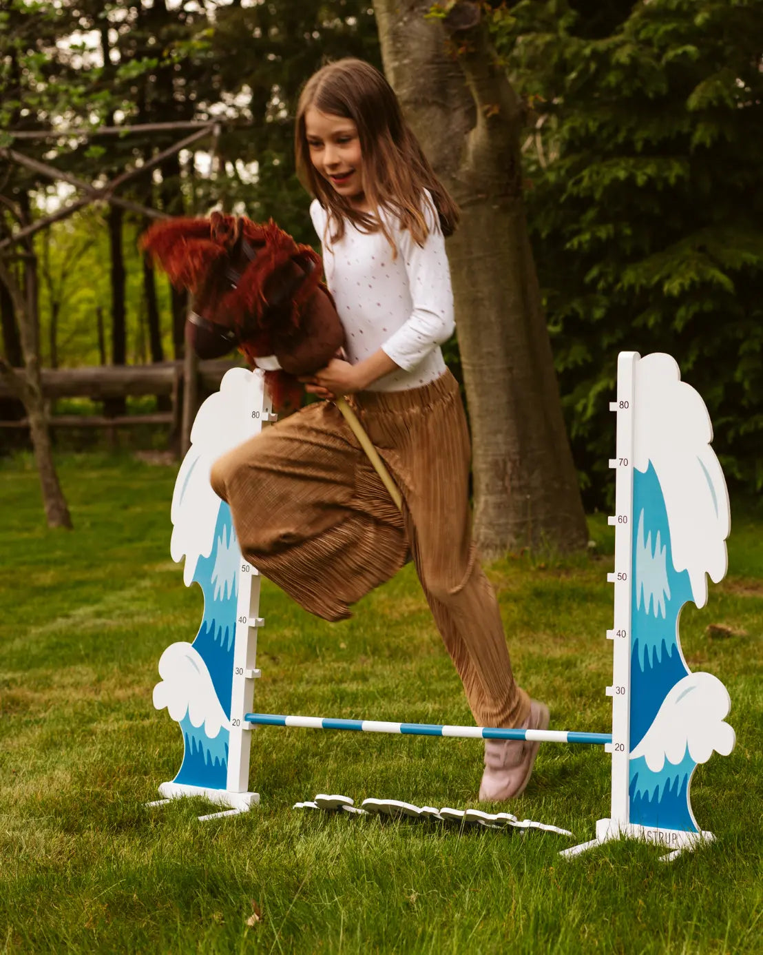 Mädchen springt mit einem Steckenpferd über das blaue byASTRUP Hobby Horsing Hindernis Wave auf einer Wiese.