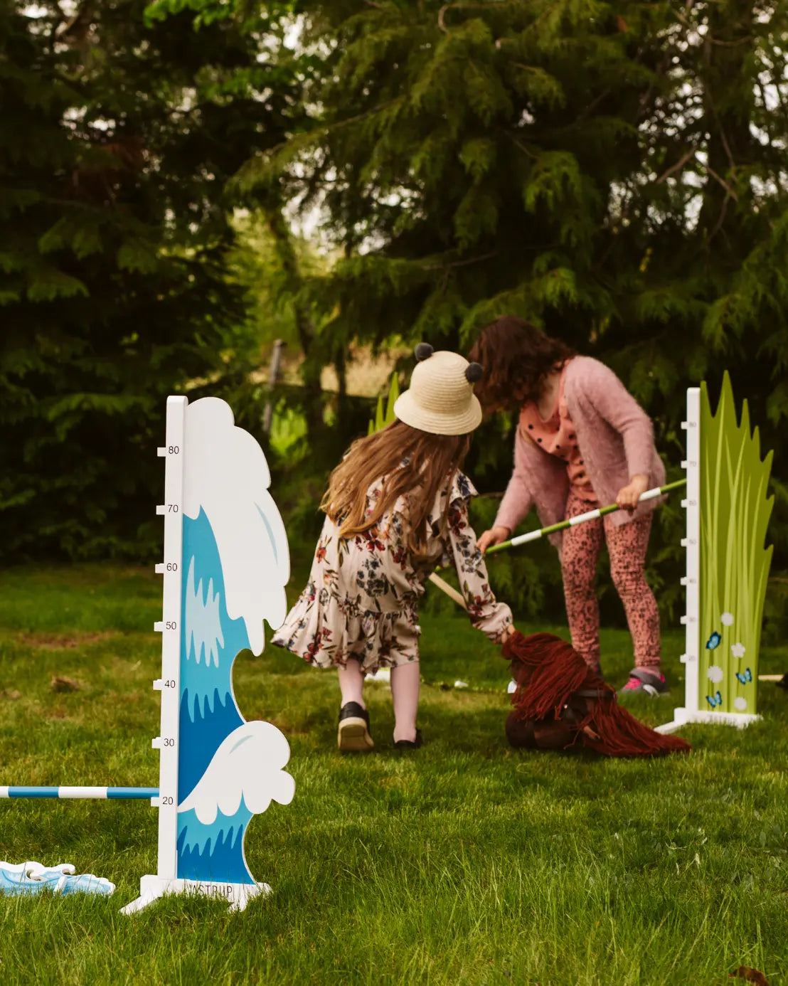 Zwei Kinder bauen das byASTRUP Hindernis für Steckenpferde im Garten auf; Fokus auf die blauen Wellen-Seitenteile.